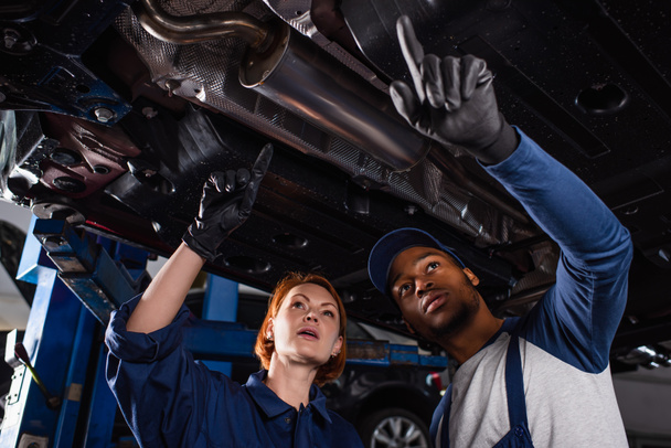 Low angle view of multiethnic mechanics in gloves pointing with fingers under car in service  - Photo, Image
