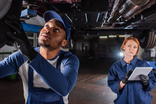African american mechanic looking at car wheel near blurred colleague with digital tablet in service  - Zdjęcie, obraz