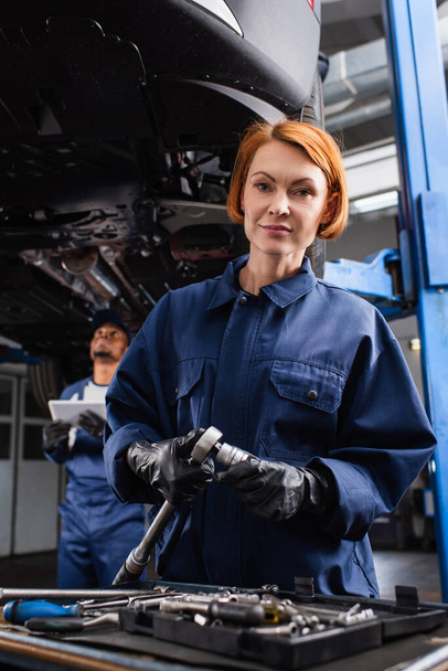 Workwoman holding wrench and looking at camera near blurred colleague and car in service  - Foto, Imagem