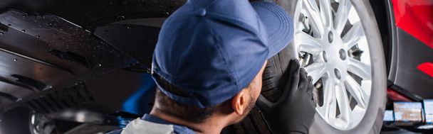 African american mechanic in cap looking at car wheel in garage, banner  - Valokuva, kuva