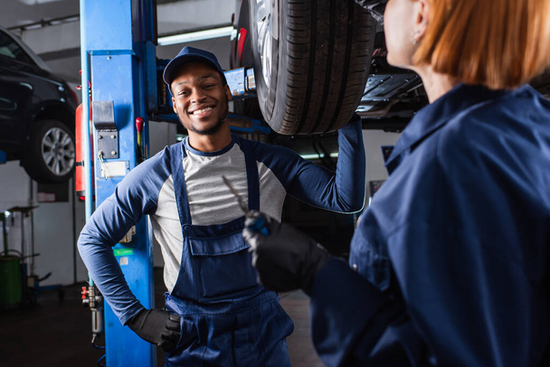 Usmívající se africký americký mechanik při pohledu na kameru v blízkosti auta a kolega v servisu  - Fotografie, Obrázek