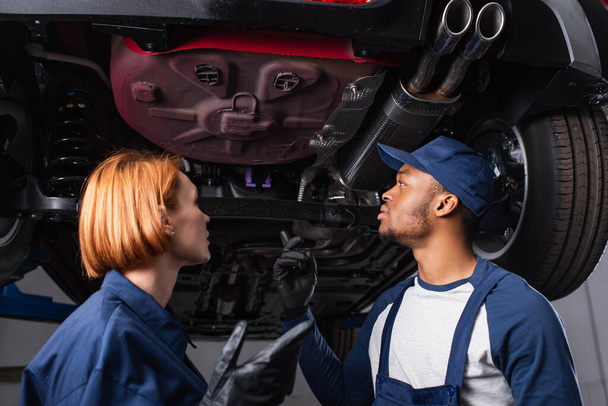 Nízký úhel pohledu multiethnic mechaniky mluví pod autem v garáži  - Fotografie, Obrázek