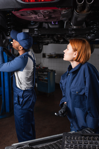 Side view of redhead mechanic holding wrench near african american colleague working with car in service  - Zdjęcie, obraz