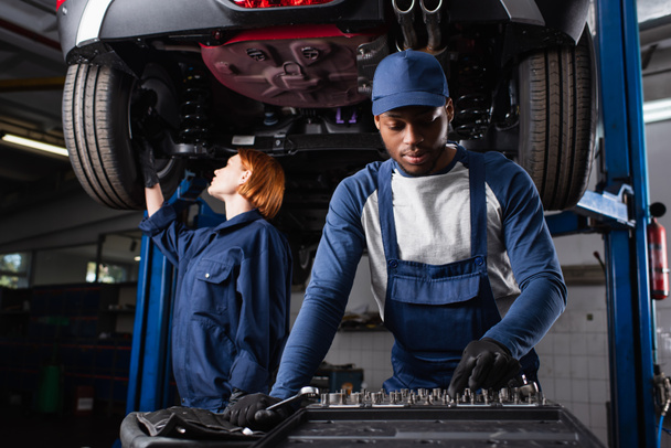 Africano americano mecánico tomando herramienta cerca colega trabajando con coche en garaje  - Foto, Imagen