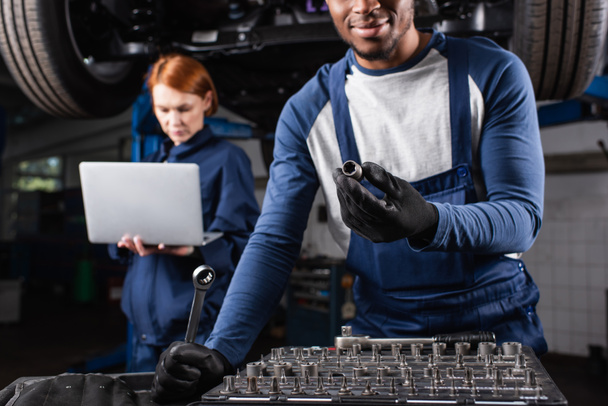 African american repairman holding tools near blurred colleague with laptop in car service  - Foto, Imagen