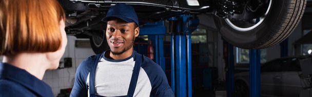 Hymyilevä afrikkalainen amerikkalainen mekaanikko katselee sumeaa kollegaa autopalvelussa, banneri  - Valokuva, kuva