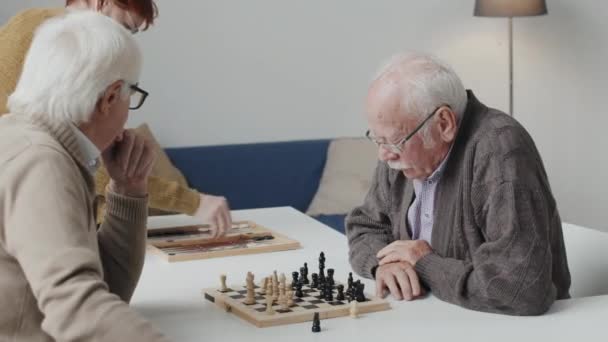 Medium shot of two thoughtful Caucasian senior men playing chess at table in nursing home - Filmati, video