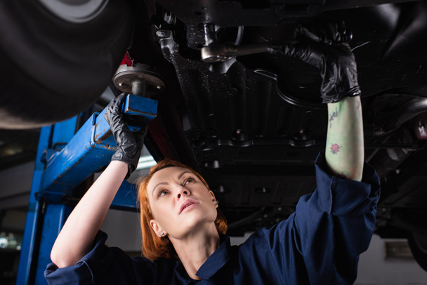 Rotschopf und tätowierter Mechaniker bei der Arbeit unter dem Auto im Dienst  - Foto, Bild