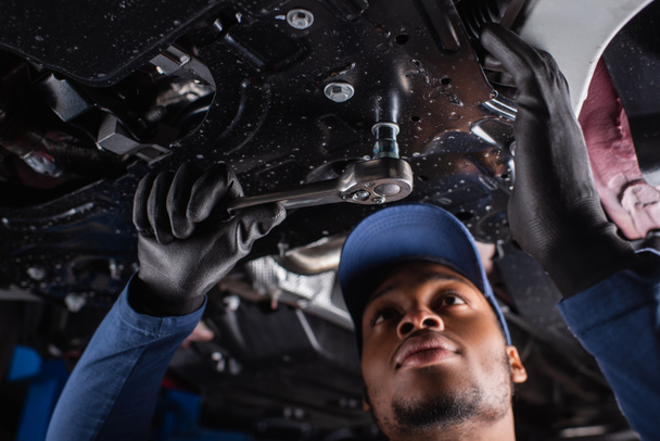 Niedrigwinkel-Ansicht von afrikanisch-amerikanischen Mechaniker in Mütze mit Schraubenschlüssel während der Arbeit im Autoservice  - Foto, Bild