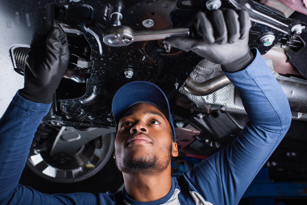 Νεαρός αφροαμερικανός μηχανικός που εργάζεται με το εργαλείο κάτω από το αυτοκίνητο στο γκαράζ  - Φωτογραφία, εικόνα