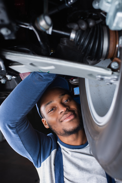 Afrykański amerykański mechanik uśmiechający się do kamery podczas inspekcji amortyzatora samochodu - Zdjęcie, obraz