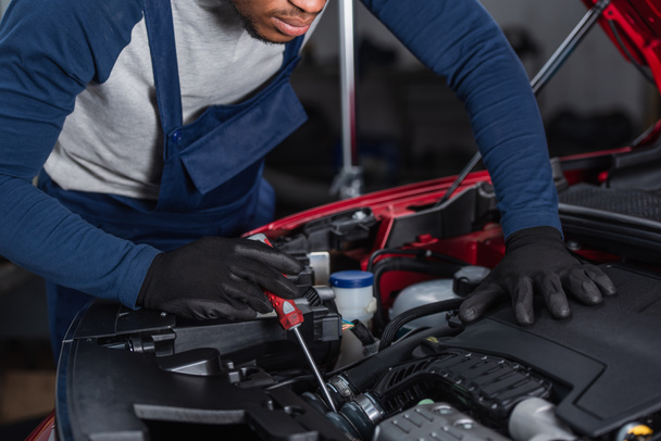 partial view of african american technician in work gloves inspecting car engine compartment with screwdriver - Foto, Bild