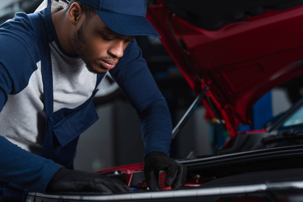 african american technician in uniform and work gloves inspecting car in workshop - Fotografie, Obrázek