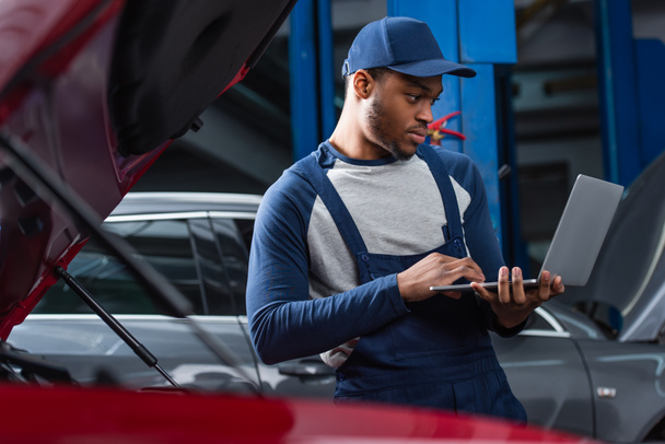 young african american foreman using laptop near blurred cars in workshop - Foto, afbeelding