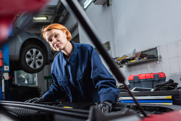 forewoman in uniform looking at camera near blurred car in workshop - Photo, Image