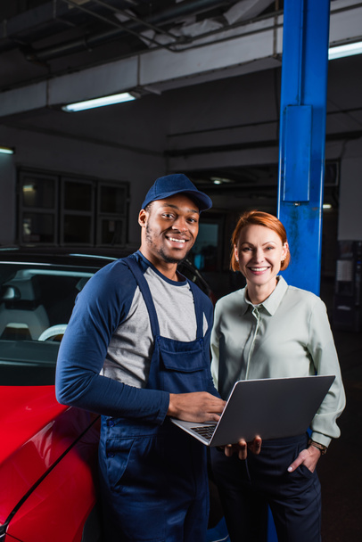 happy client and african american mechanic with laptop smiling near car in workshop - 写真・画像
