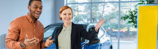 usmívající se prodejce aut ukazuje rukou téměř ohromený africký americký klient, banner - Fotografie, Obrázek