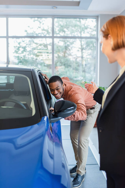 difuminado concesionario de coches apuntando con la mano cerca de hombre afroamericano feliz mirando dentro de coche en sala de exposición - Foto, Imagen