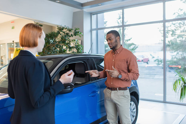 Afrikaans amerikaanse man wijzend met de handen op de auto, terwijl op zoek naar wazig auto dealer - Foto, afbeelding