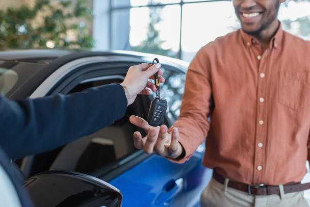 przycięty widok dealera samochodów dając klucz do afrykańskiego amerykańskiego klienta uśmiechającego się na rozmytym tle - Zdjęcie, obraz