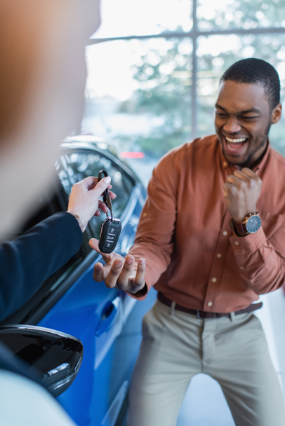 ενθουσιασμένος αφρικανός Αμερικανός άνθρωπος δείχνει την επιτυχία χειρονομία, ενώ λαμβάνοντας το κλειδί από θολή έμπορος αυτοκινήτων - Φωτογραφία, εικόνα
