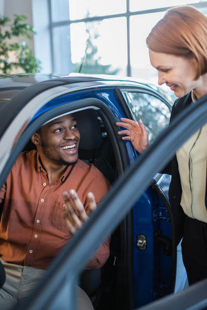 hombre afroamericano feliz hablando con distribuidor de coche borroso mientras está sentado en auto  - Foto, Imagen