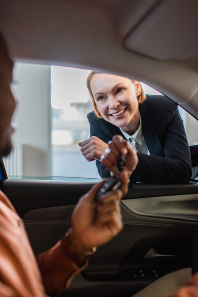 cheerful car dealer giving key to blurred african american buyer sitting in car - Photo, Image