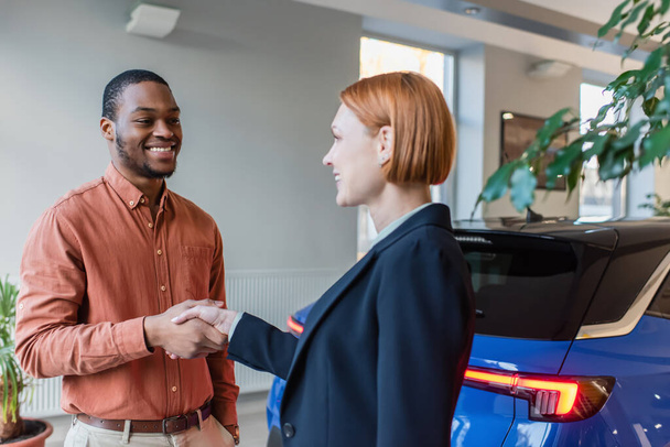 happy african american man and car dealer shaking hands near car  - Фото, изображение