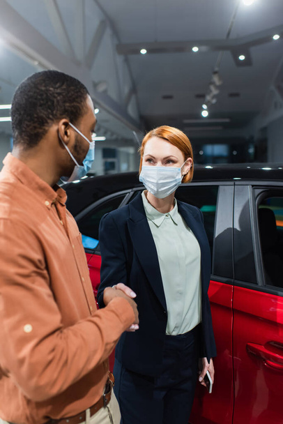 car dealer and african american client in medical masks shaking hands near car - Foto, Bild