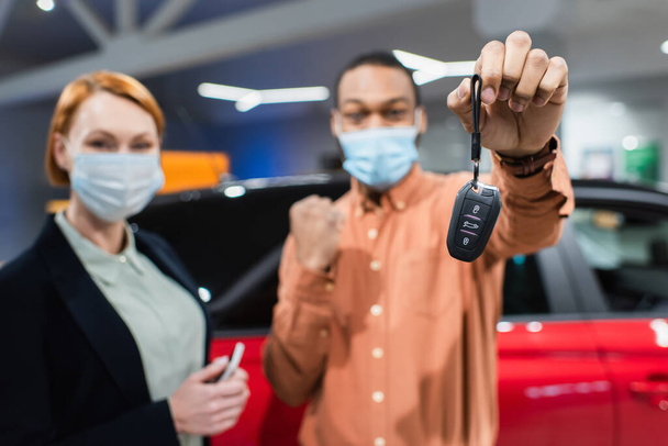 selektivní zaměření klíče v ruce rozmazané africký Američan muž v lékařské masce ukazuje úspěch gesto v blízkosti prodejce aut - Fotografie, Obrázek