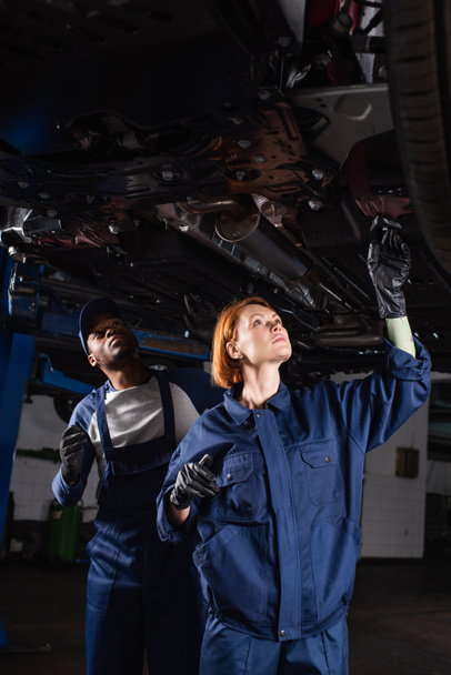travailleurs interraciaux du service automobile inspectant le bas de l'auto soulevée en atelier - Photo, image