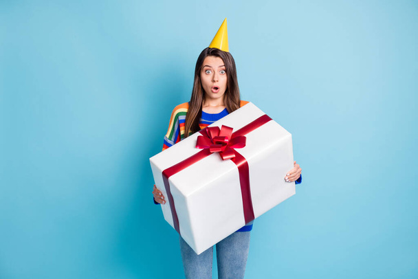 Photo portrait of shocked woman keeping big present box wearing yellow cap on party isolated on bright blue color background - 写真・画像