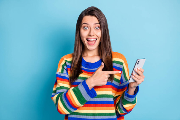 Photo portrait of girl amazed pointing finger smartphone smiling using app isolated on vibrant blue color background - 写真・画像