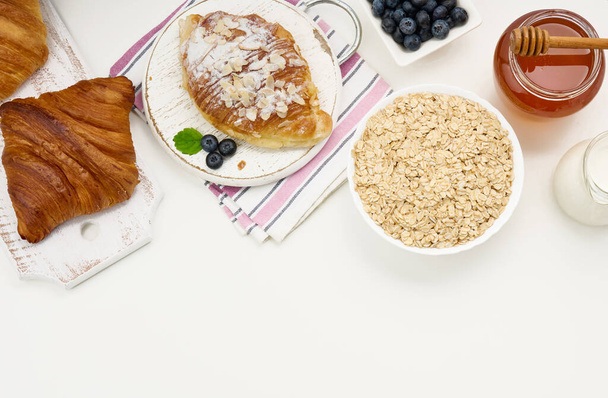 Сырая овсянка в белой керамической тарелке, черника, мед на белом столе, завтрак. вид сверху - Фото, изображение