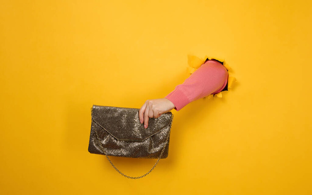 mão feminina está segurando um pequeno saco de embreagem dourada com uma corrente de metal em um fundo amarelo. Parte do corpo saindo de um buraco rasgado em um fundo de papel - Foto, Imagem