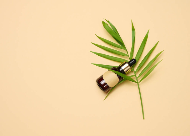 brown glass bottle with a pipette and with blank paper label stands on a brown background. Container for cosmetics, serum and oil, palm leaf  - Photo, Image