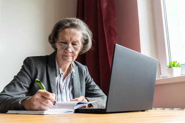pääaihe on epätarkka, vanhempi vanha nainen istuu vieressä pöydällä, kotitoimisto verkossa kannettava tietokone työ liiketoiminnan etäisyys freelancer kirjoittaa kynä, tehdä muistiinpanoja - Valokuva, kuva