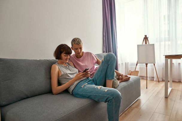 Lesbian girls watch on smartphone on sofa at home - 写真・画像