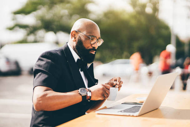 Selective focus on a stylish bearded gentleman sitting outdoors using a notebook and holding a cup. A black man with glasses on a table, looking at the notebook screen and stirring a cup of coffee - Fotoğraf, Görsel