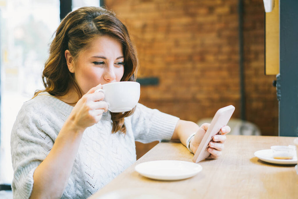 Young woman with cup of coffee in hand messaging, reading, texting, watching video, scrolling social networks, making notes, online shopping on phone while sitting at table in modern cafe - Zdjęcie, obraz
