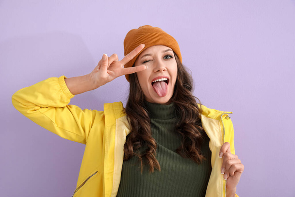 Emotional woman in raincoat showing tongue and peace gesture on color background - Photo, Image