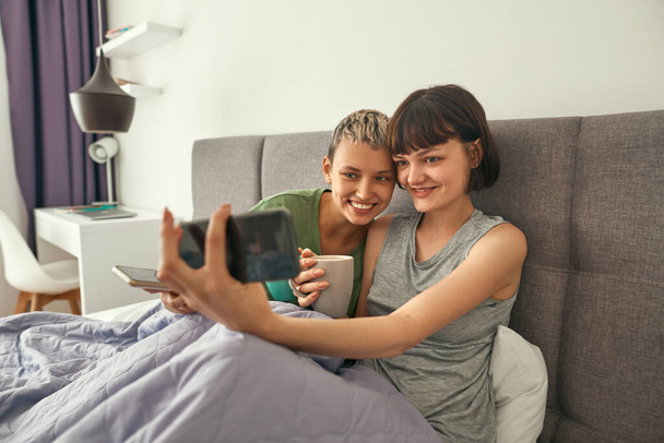 Lesbians with tea take selfie on smartphone on bed - Photo, image