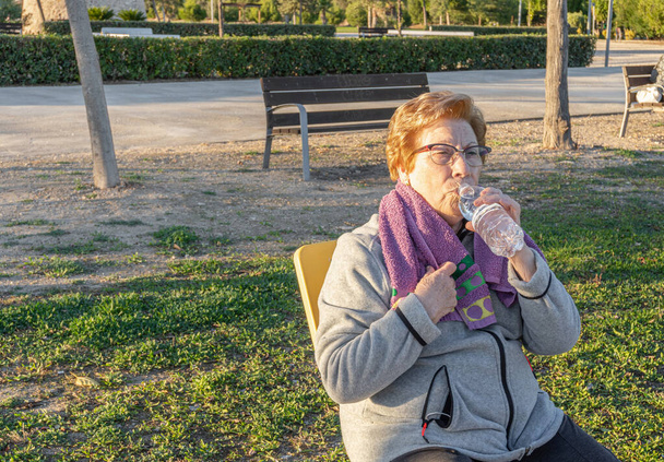 Mujer mayor bebe agua de la botella después de hacer ejercicio en un parque al atardecer - Foto, Imagen