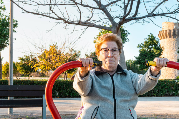 Ηλικιωμένη γυναίκα που γυμνάζεται σε βιο-υγιεινά μηχανήματα σε ένα πάρκο το ηλιοβασίλεμα. - Φωτογραφία, εικόνα
