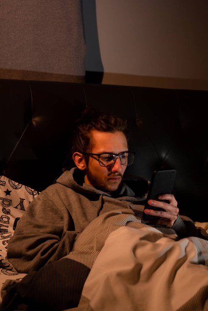 Молодий чоловік, залежний від соціальних медіа, перевіряє телефон, перш ніж іти спати, пристрасть, соціальні мережі, після темної концепції. - Фото, зображення