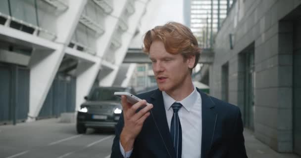 Concentrated businessman recording voice message standing outdoors at business district. Emotional red-haired male entrepreneur talking on mobile phone speakerphone, conducting aggressive negotiations - Footage, Video