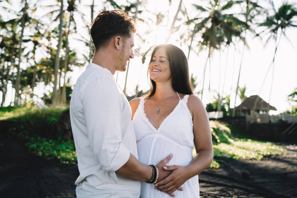 Cheerful pregnant couple standing close and caressing gently while spending summer holidays together in tropical garden with palms under bright sunlight - Фото, зображення