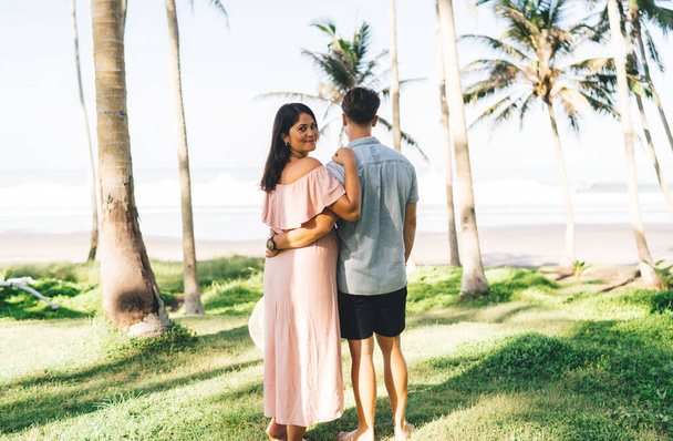 Back view loving young couple in stylish summer outfits hugging and admiring tranquil seascape while spending romantic honeymoon in exotic resort - Foto, Imagem