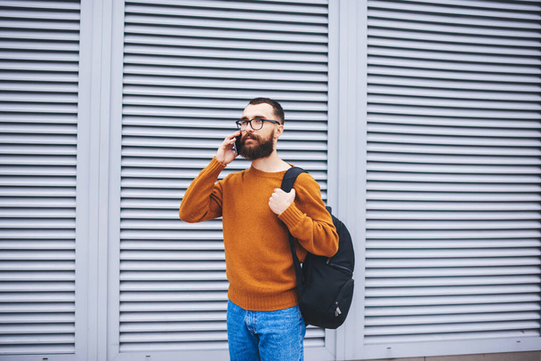 Thoughtful bearded male wearing casual clothes looking away standing on street near wall talking on mobile phone having free time - Photo, image