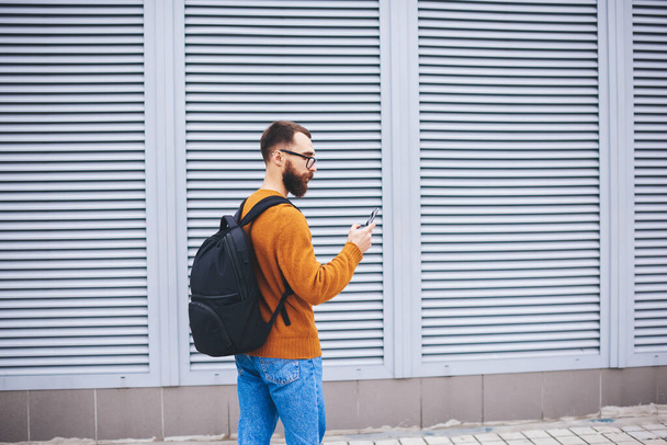 Boční pohled na mladé vousaté hipster v chytré ležérní nosit brýle a černý batoh čtení textové zprávy na mobilním telefonu vedle moderní budovy - Fotografie, Obrázek